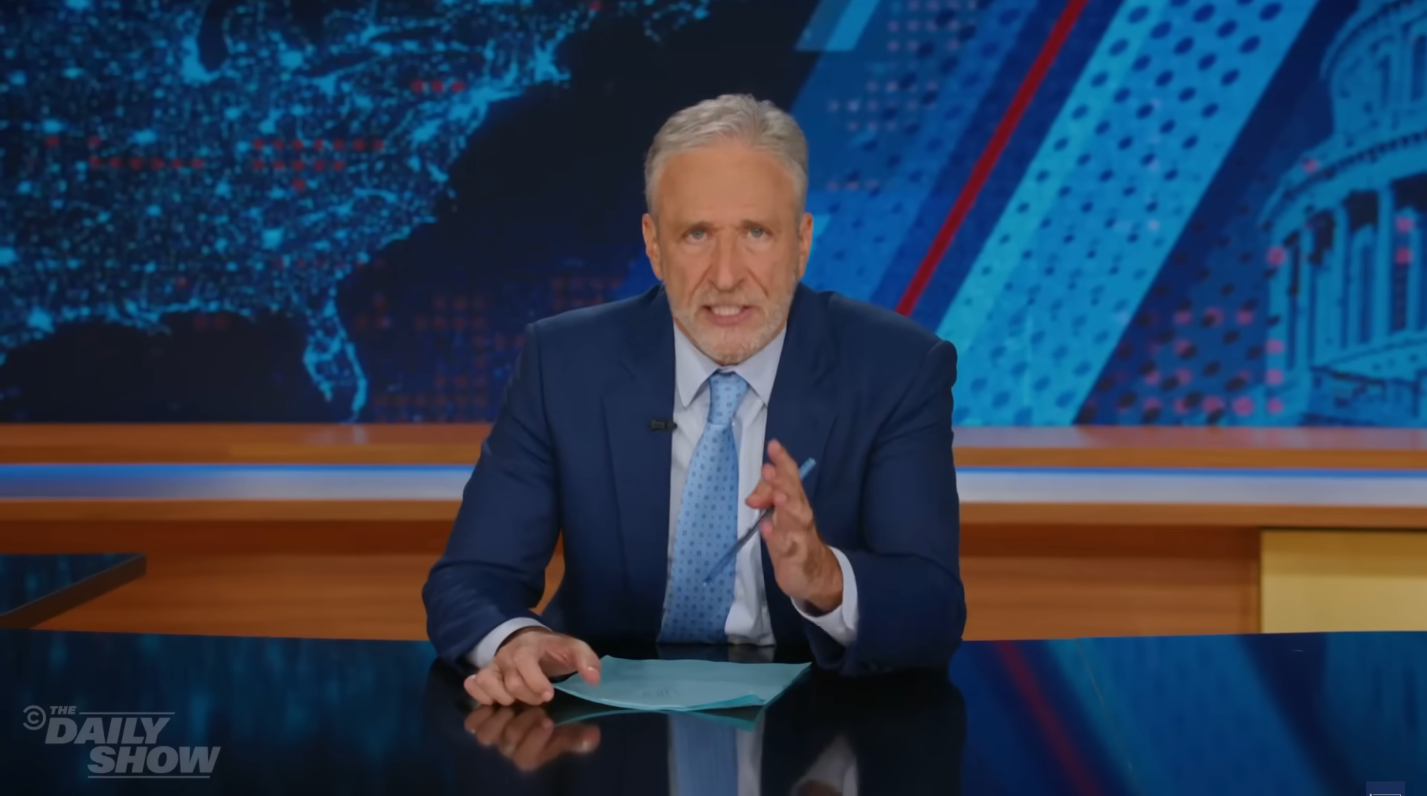 A news anchor in a suit sits at a desk with a serious expression, holding papers. A map and patterns are in the background