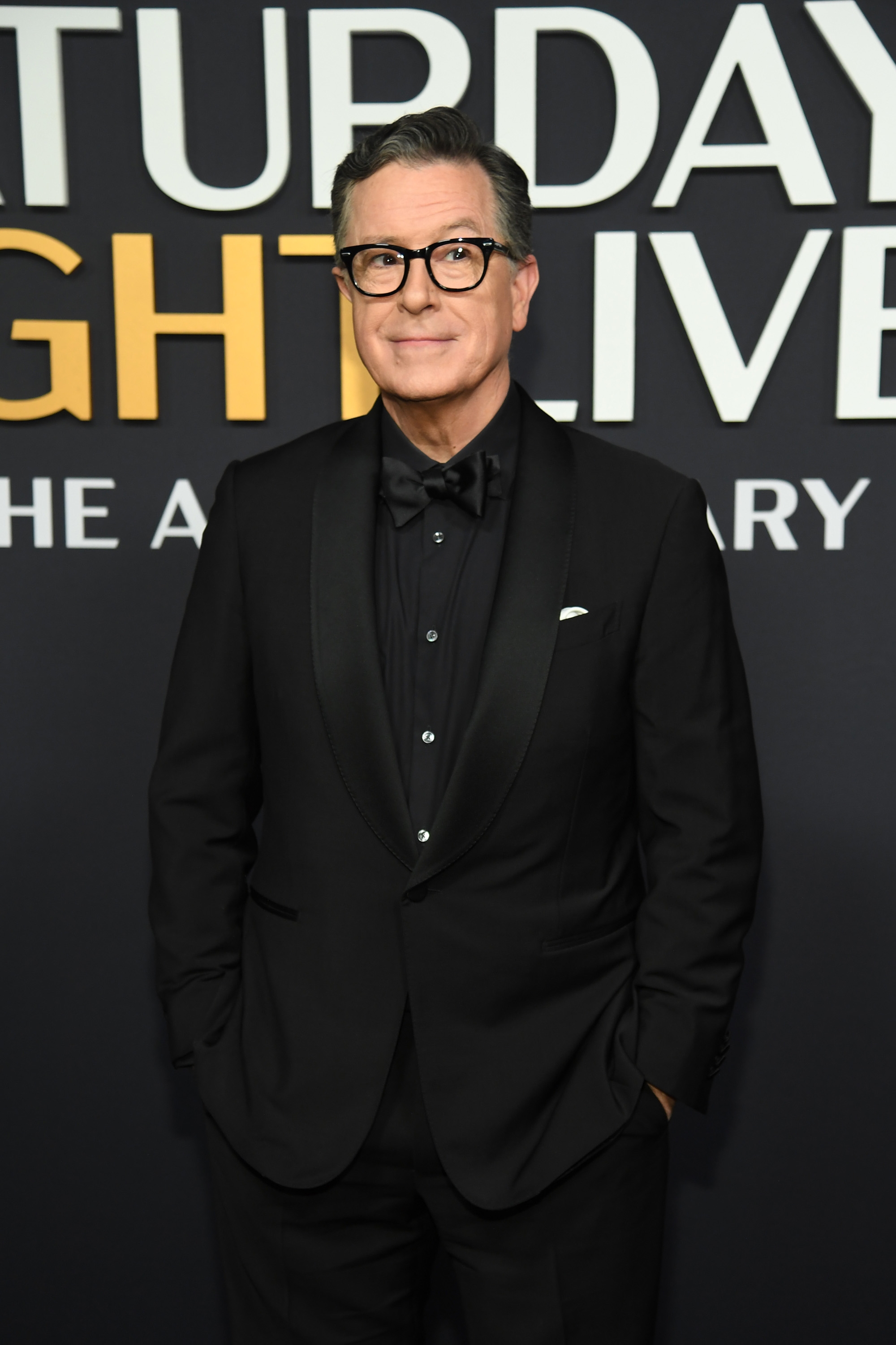 Person in a formal black suit and bow tie standing in front of a &quot;Saturday Night Live&quot; backdrop