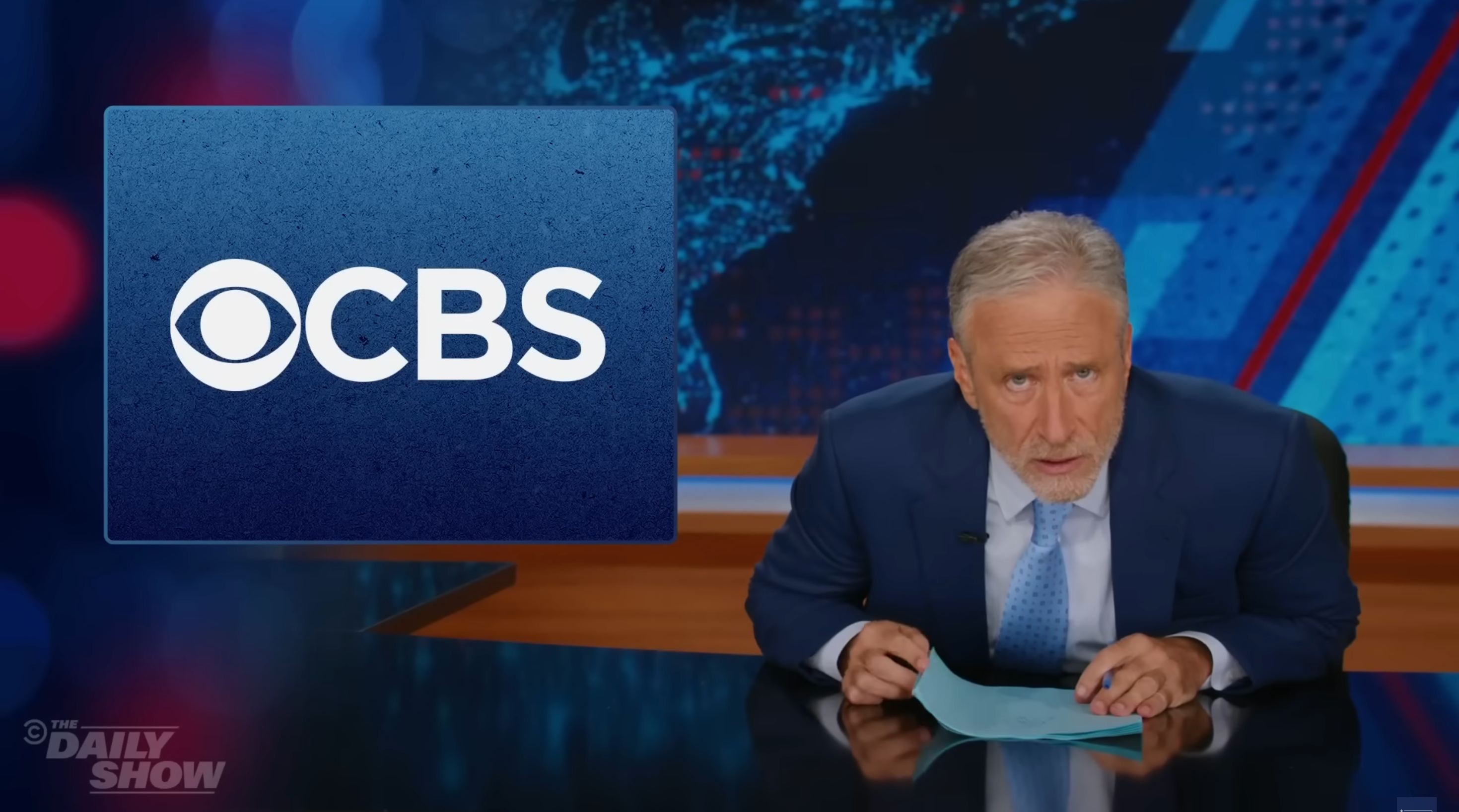 A person at a news desk looks seriously at the camera; a CBS logo is displayed on a screen beside them