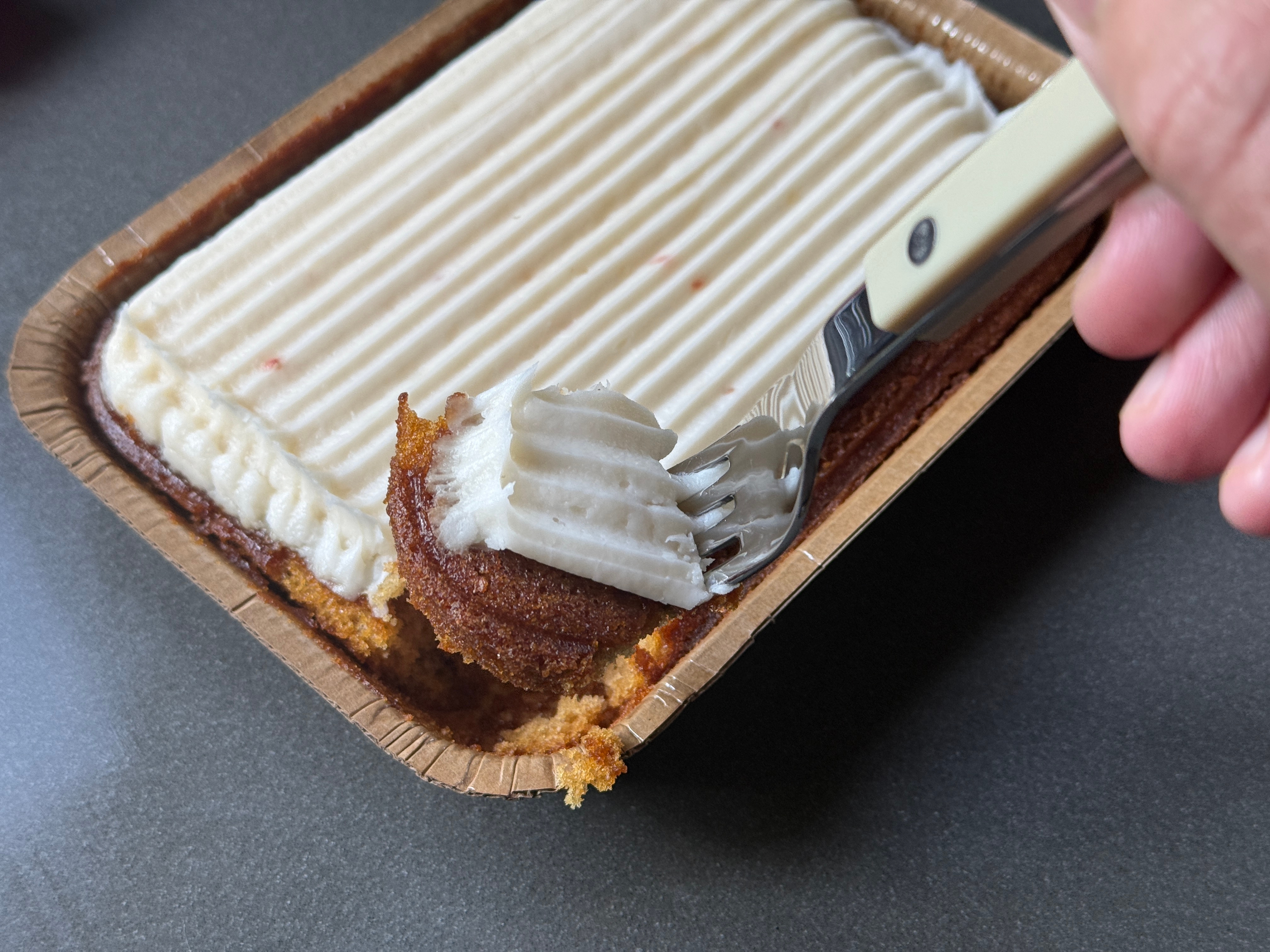 A person holds a fork taking a piece from a small rectangular cake covered in frosting
