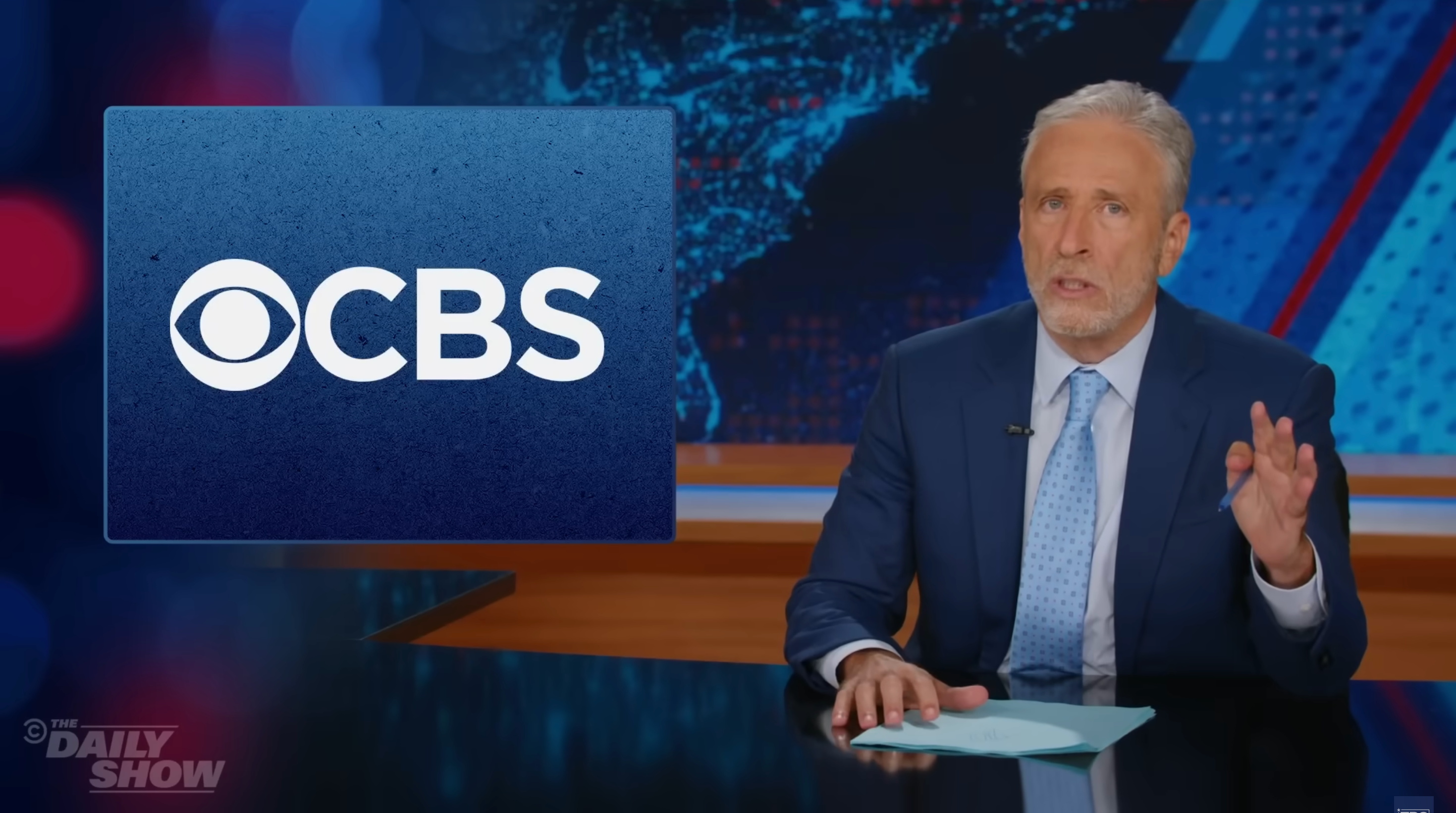 A well-known TV host wearing a suit sits at a news desk with a &quot;CBS&quot; logo displayed on a screen beside him
