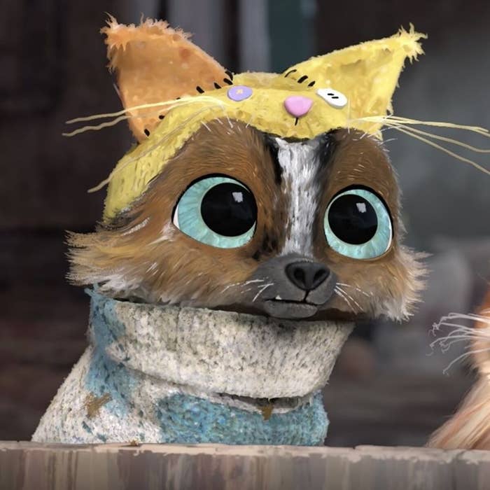 Un personaje animado con aspecto de gato lleva un gorrito amarillo y bufanda celeste, con ojos grandes y expresivos, mirando al frente