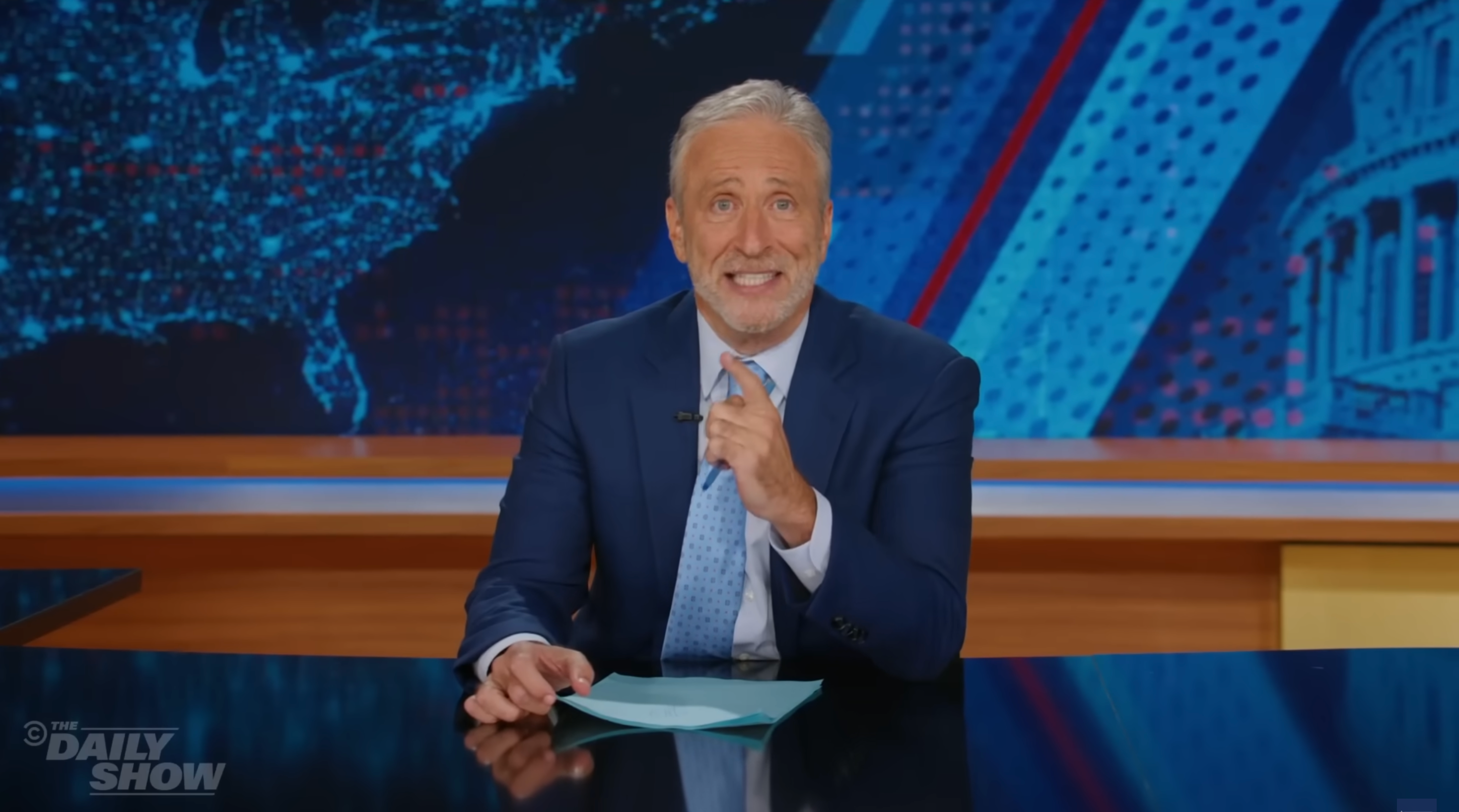 A man in a suit sits at a news desk, gesturing towards the camera, with a map and columns in the background