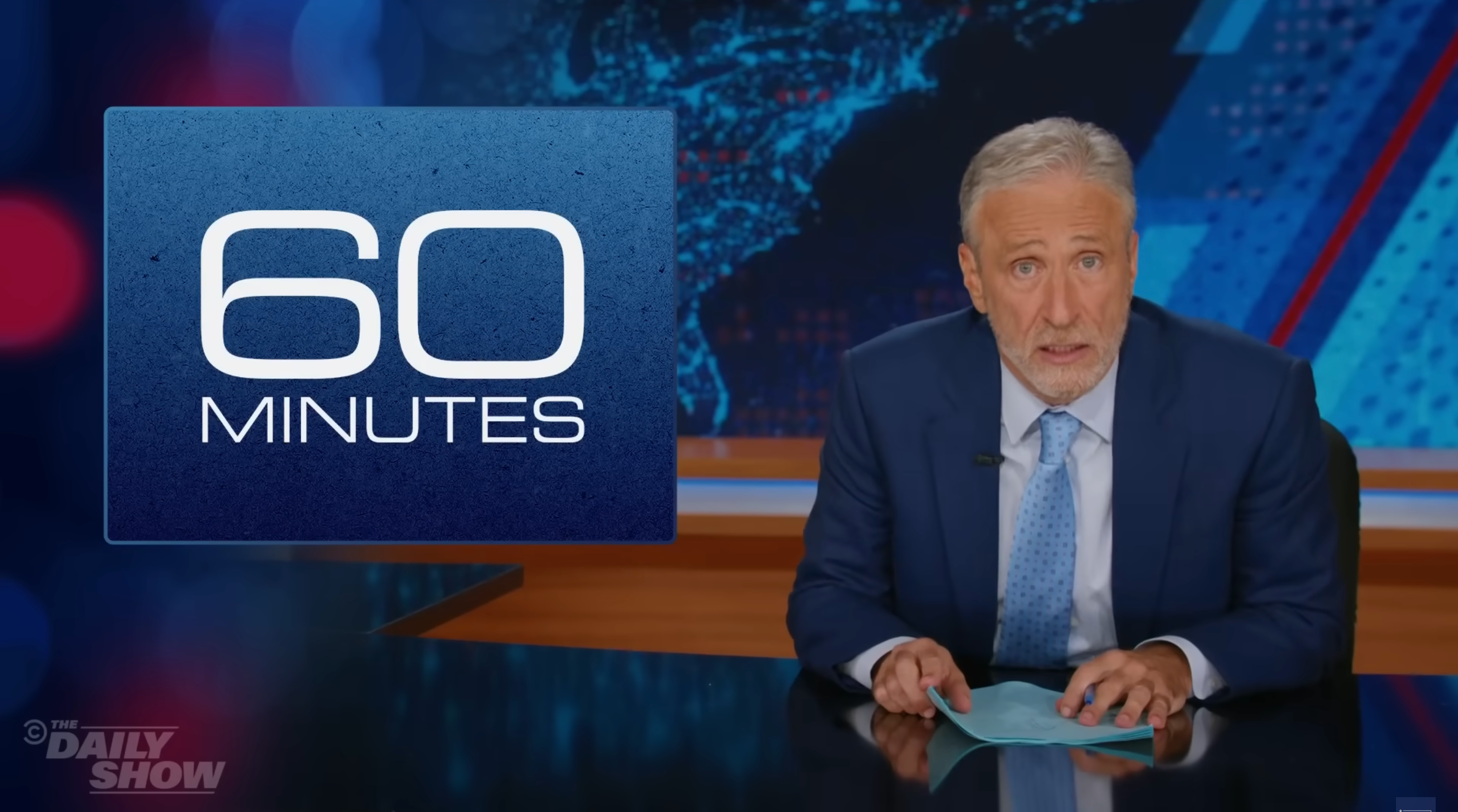 A man in a suit sits at a news desk with a &quot;60 Minutes&quot; logo in the background