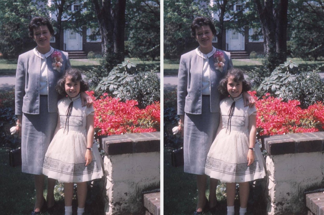 A woman and a girl pose outdoors; the woman wears a suit with a floral corsage, and the girl wears a dress with a ribbon tie, standing by flowers
