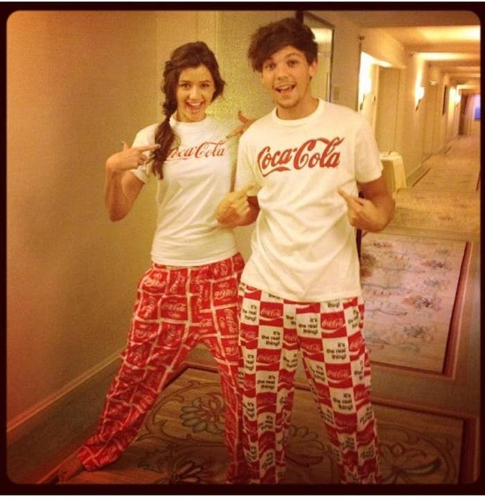 Dos personas en un pasillo, vistiendo camisetas blancas y pantalones con logotipos de Coca-Cola. Ambos sonríen y señalan sus camisetas