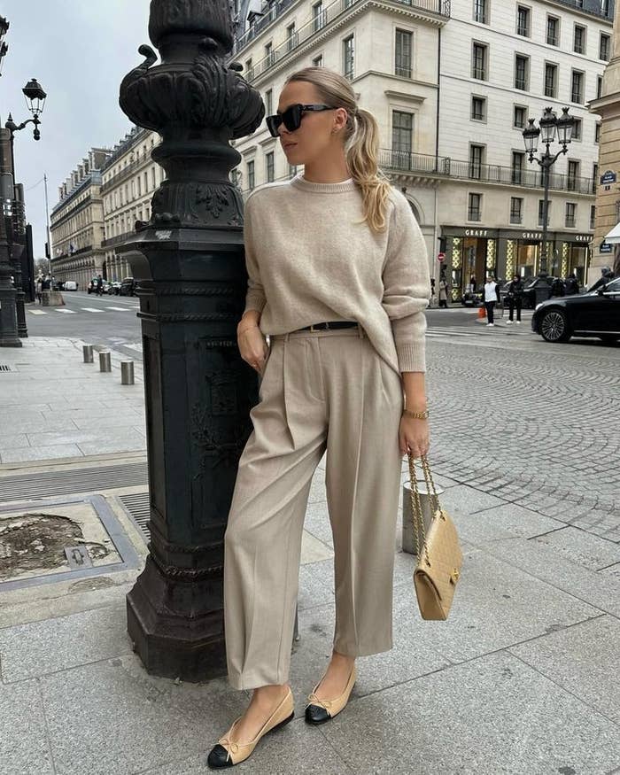 Mujer en una calle elegante, con suéter suelto y pantalones de vestir. Lleva gafas de sol, bolso de mano y bailarinas bicolor