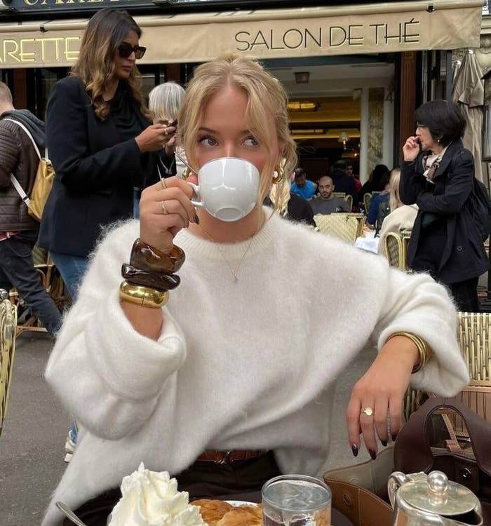 Persona en café parisino, bebiendo té. Lleva suéter holgado y pulseras grandes. Mesa con croissant, crema y tetera. Ambiente relajado
