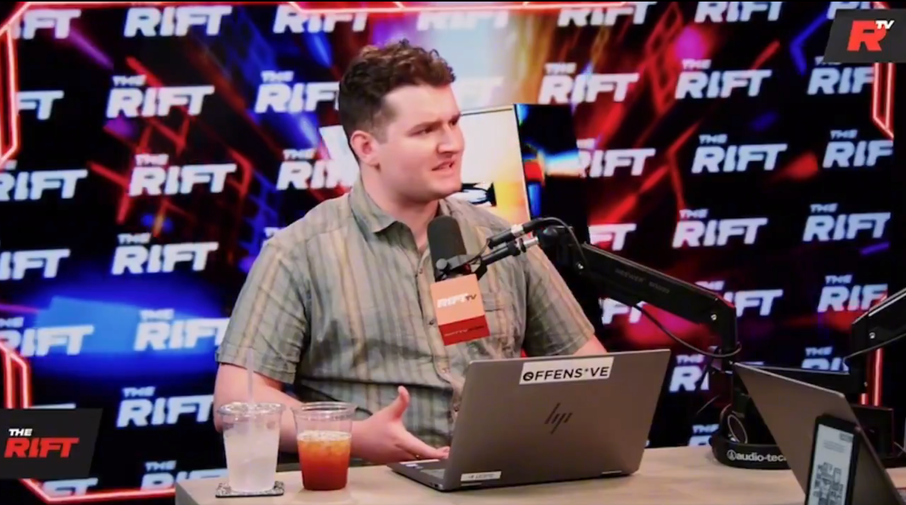 Person speaking into a microphone in a podcast studio setting with &quot;The Rift&quot; branding in the background
