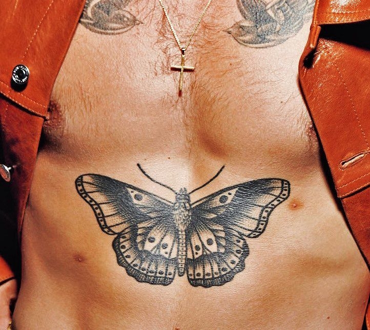 Torso masculino con una cadena y crucifijo, tatuaje de mariposa en el estómago. Lleva chaqueta parcialmente abierta