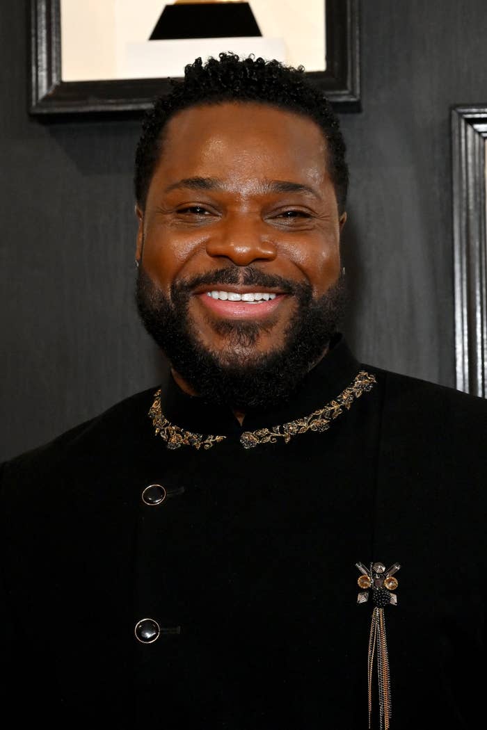 Malcolm Jamal Warner smiling on the red carpet, wearing an elegant suit with intricate embellishments on the collar and lapel