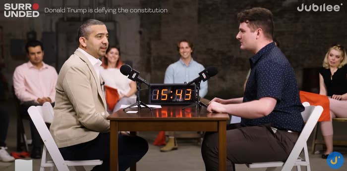 Two people seated at a table debate with a timer, surrounded by an audience. Text reads, &quot;Donald Trump is defying the constitution.&quot;