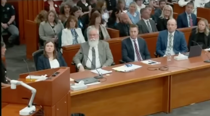 Courtroom scene with attorneys seated at a table, facing the judge. The courtroom is filled with attendees