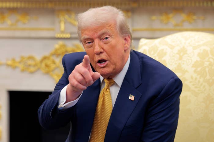 A person in a suit points forward with a serious expression while seated in an ornate room
