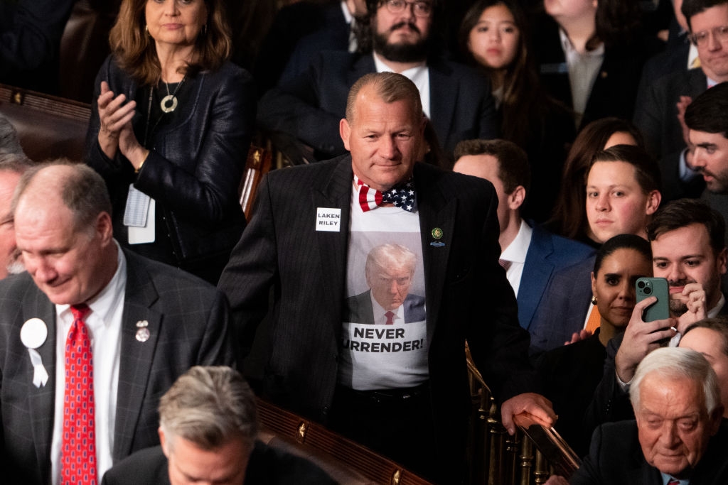 Man in a printed shirt with Trump&#x27;s image and &quot;Never Surrender&quot; slogan at a crowded event, standing among seated attendees