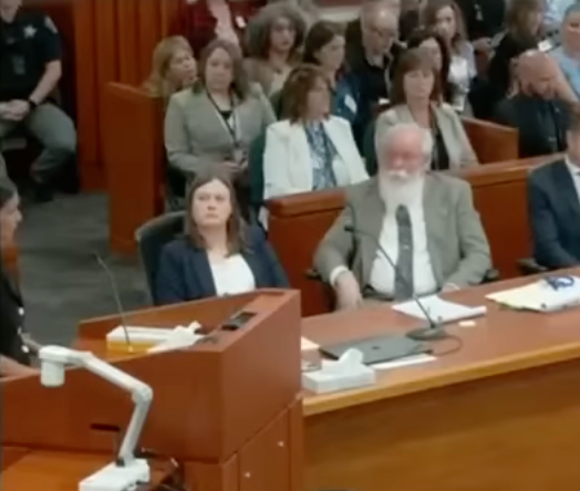 Courtroom scene showing a diverse group of people, including an older man with a white beard, seated at a table listening attentively