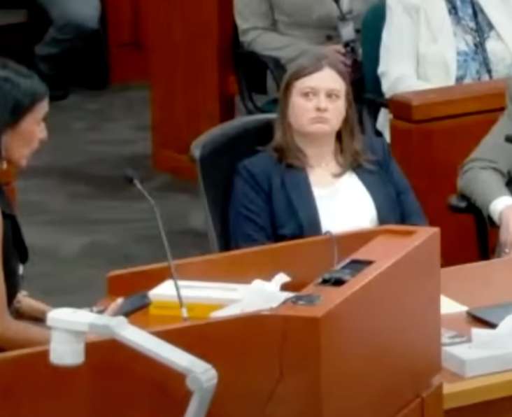 A courtroom setting with a focused woman in a blazer listening intently during a testimony