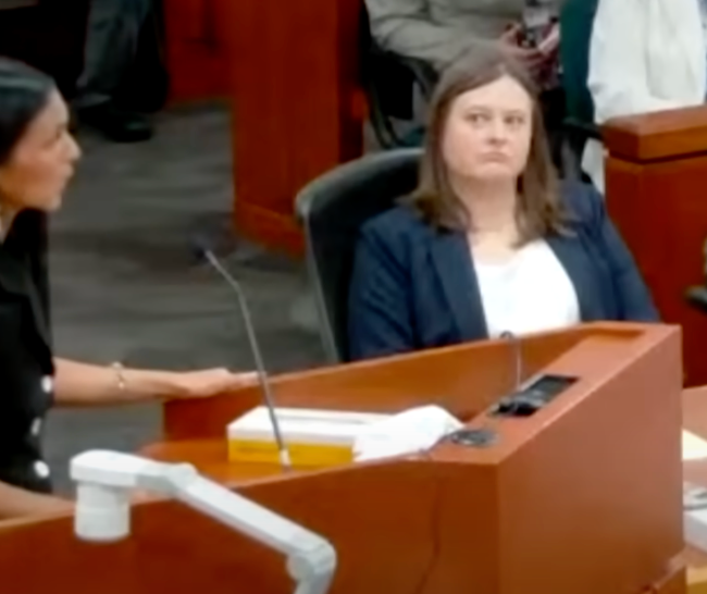 Courtroom scene with two women seated at a table, one addressing the court and the other listening attentively