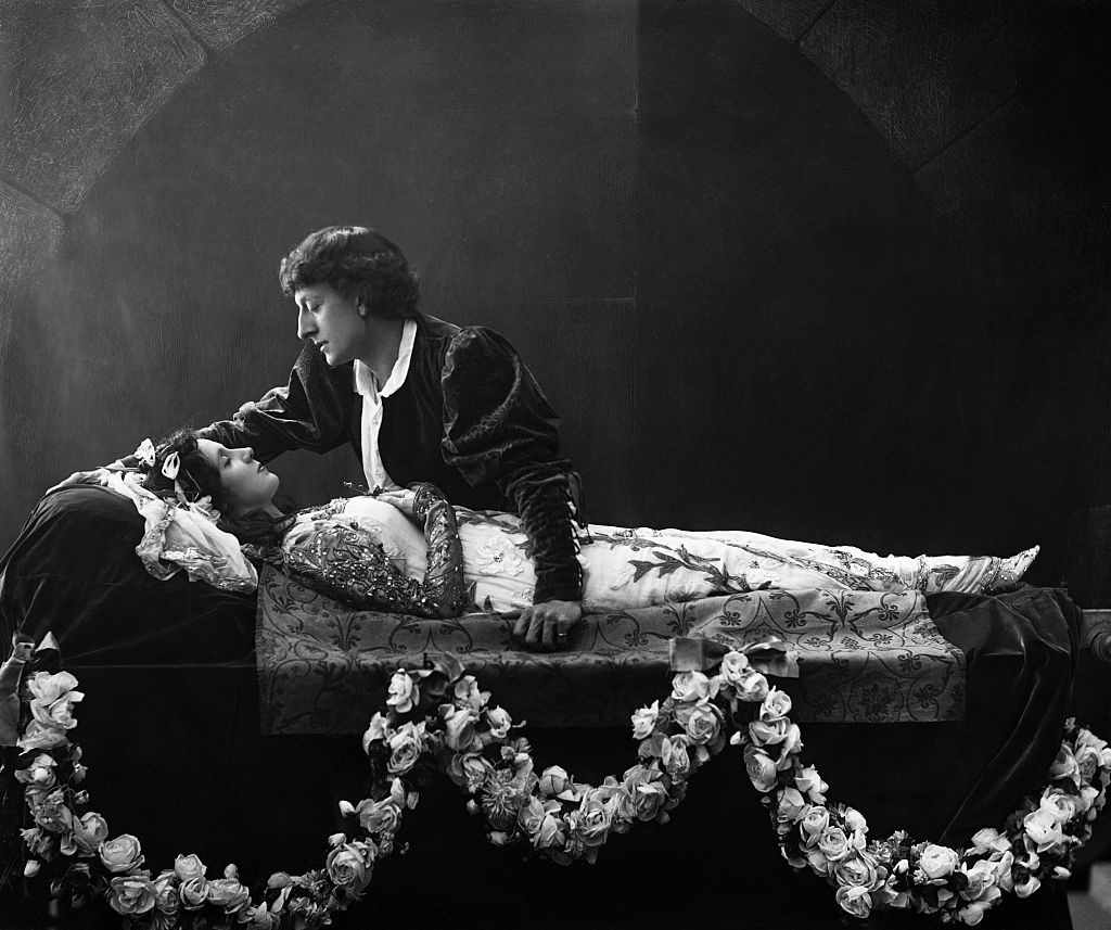 Theatrical scene with two actors in Shakespearean costumes, one lying down with eyes closed, the other leaning over in a dramatic pose