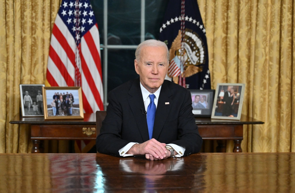 A person in a suit is seated at a desk in an official setting with flags and framed photos in the background