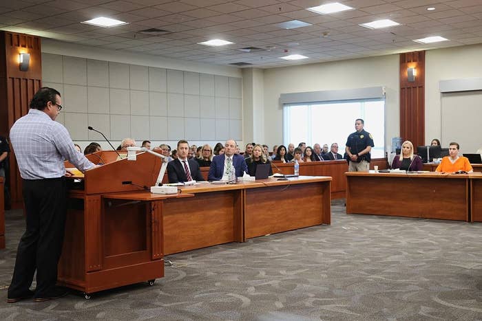 A courtroom scene with a full audience. A person speaks at a podium while others are seated at tables, listening attentively to the proceedings