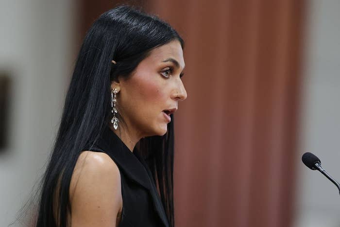 Person speaking at a podium, wearing a stylish black sleeveless outfit and long earrings, during a public or formal event