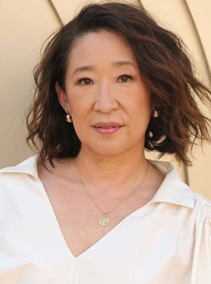 Person with wavy hair in a stylish white blouse, featuring pearl earrings and a delicate necklace, poses against a simple background