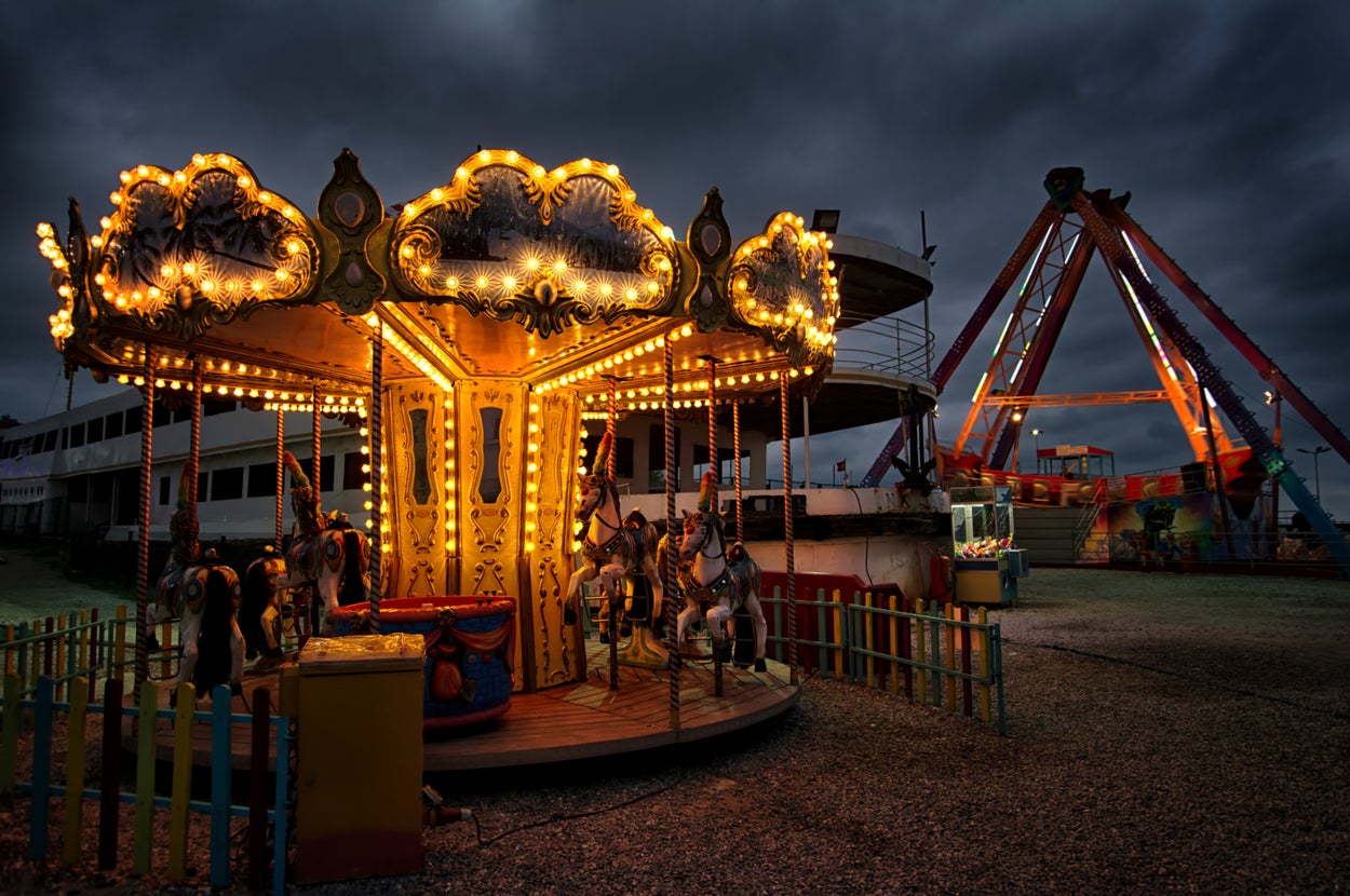 Lit carousel ride at an amusement park during nighttime, with a ship-style ride in the background under a cloudy sky