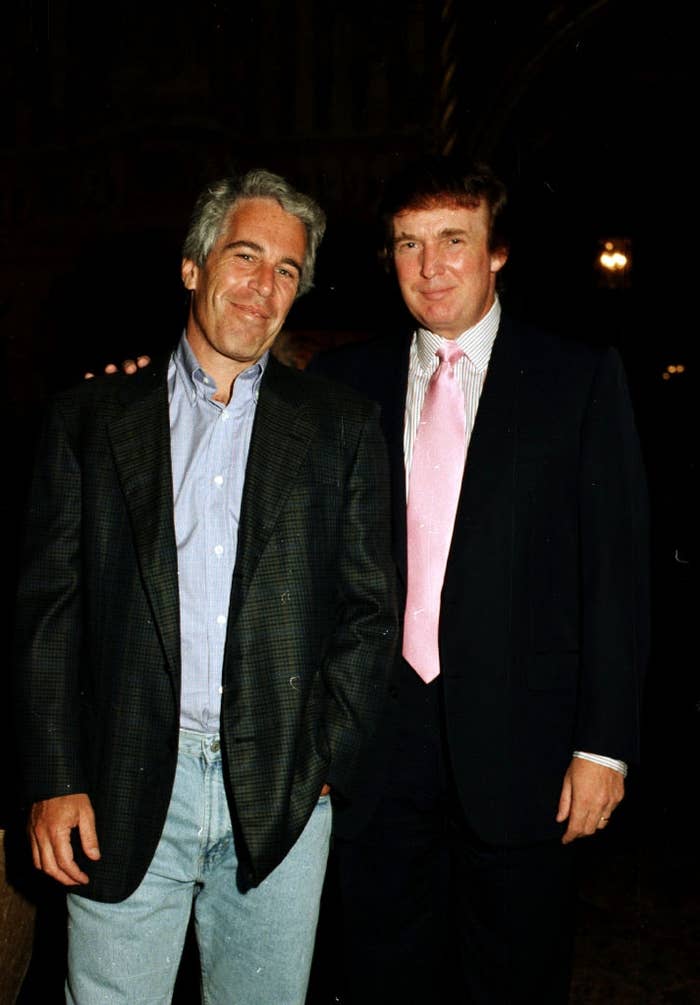 Two men in suits at a formal event. One wears a casual shirt and blazer, the other a suit with a bright tie. They stand close, smiling for the camera