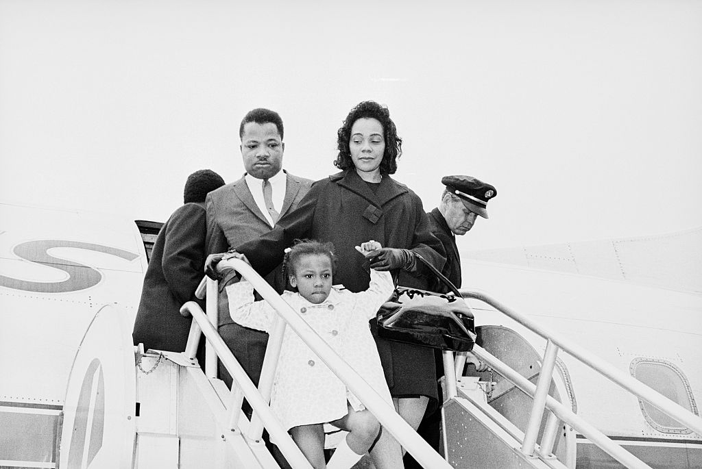A family descends airplane stairs. The man and woman are formally dressed, with a child in a light coat. An airline staff member assists them