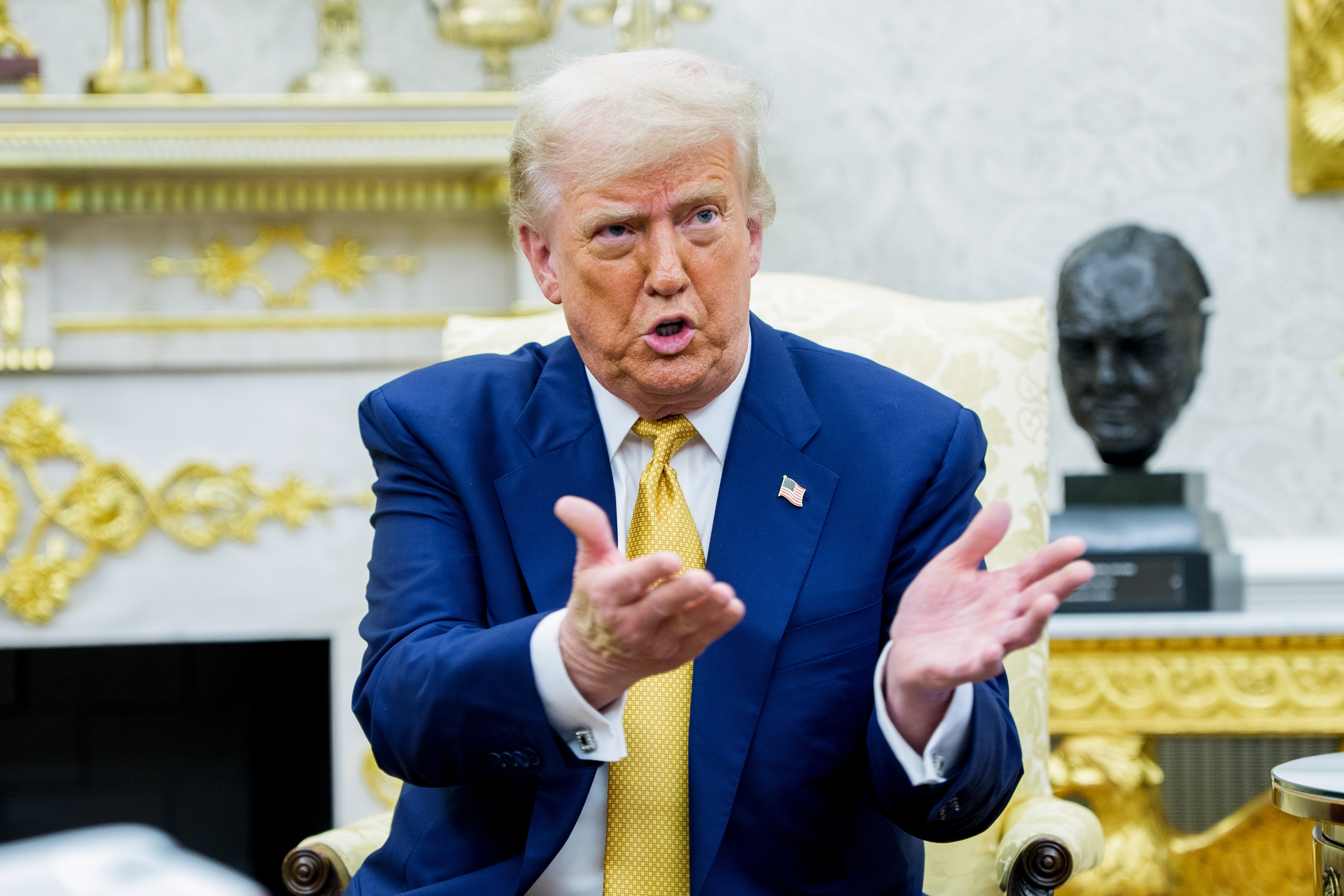 A person in a suit gestures while speaking, sitting in a formal setting with ornate decor and a sculpture in the background