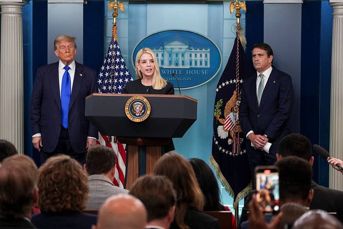 A press briefing at the White House podium with three people, including a woman speaking. The audience is taking photos and notes