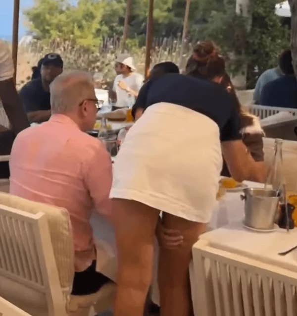 A person appears to be helping another person at a restaurant, reaching over a table set for a meal. Other diners are visible in the background