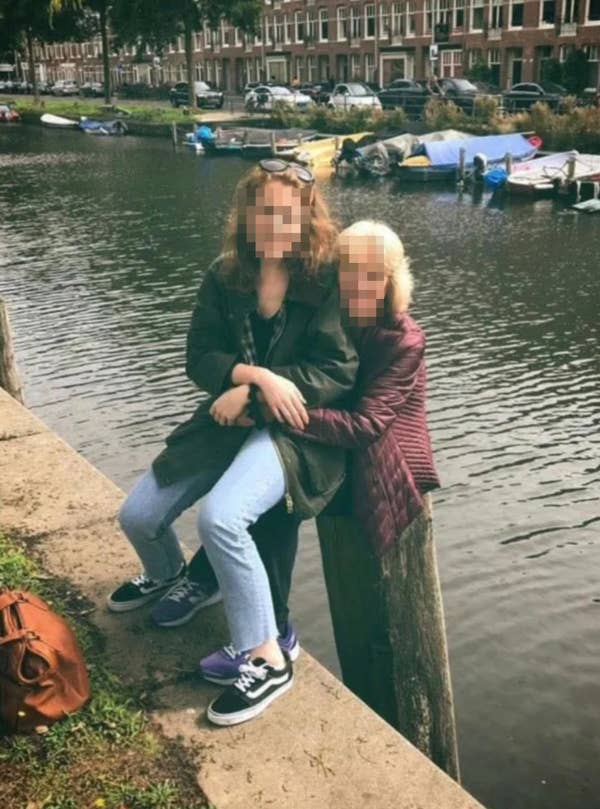 Two people smiling, sitting on a wooden post by a canal with boats in the background