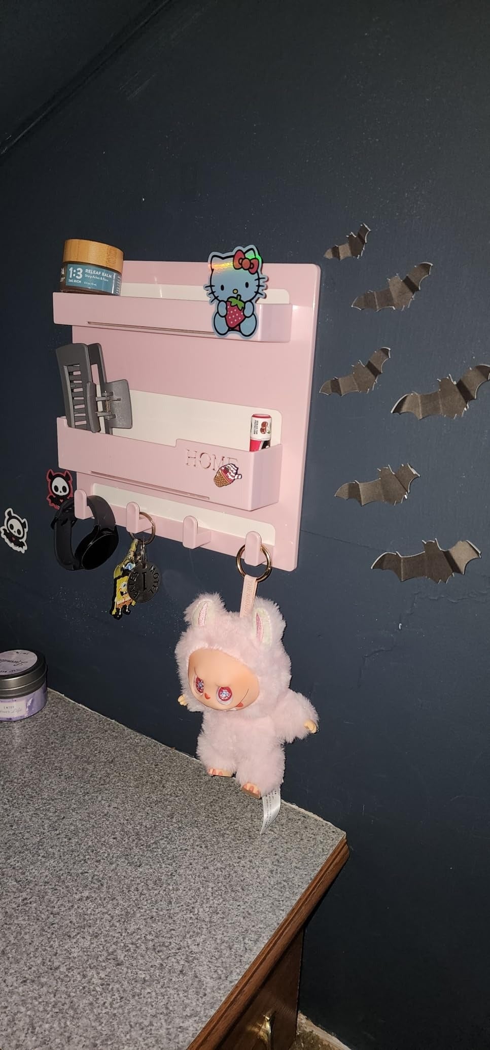Wall-mounted organizer with Hello Kitty decor, hanging plush toy, grooming products, and bats connected  the partition  supra  a granite countertop