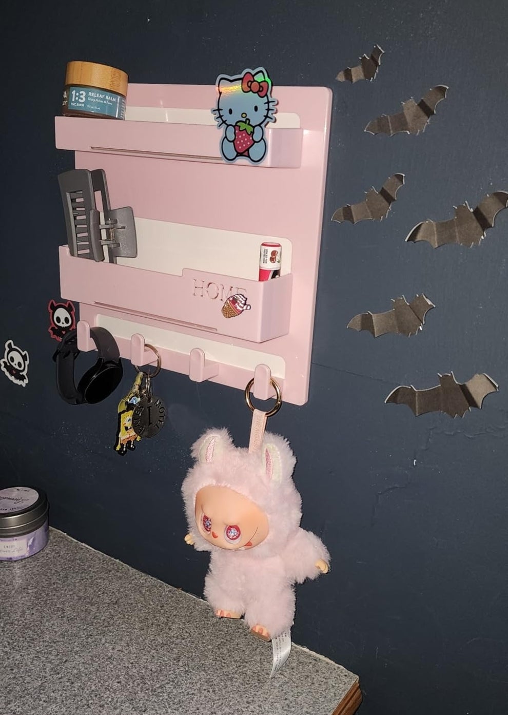 Wall-mounted organizer with Hello Kitty decor, hanging plush toy, grooming products, and bats on the wall above a granite countertop