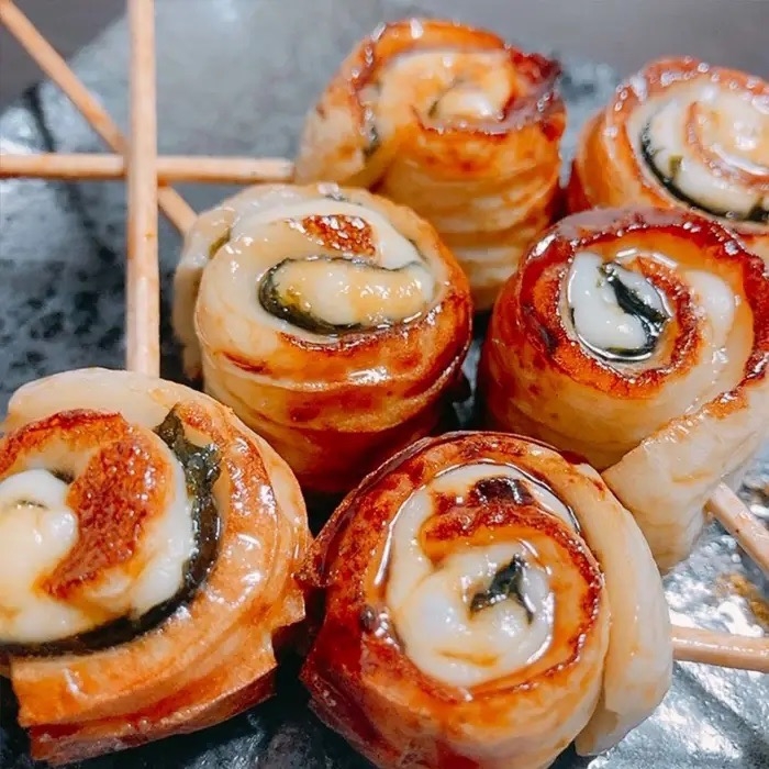 焼き竹輪の串刺しが黒い皿に盛られている。具材が巻かれて焼かれている様子が見える。