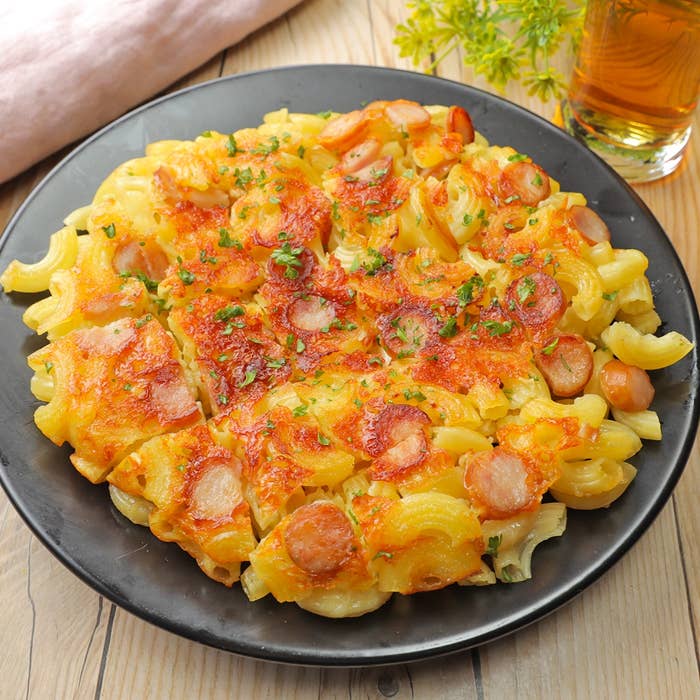 フライパンで焼いたマカロニチーズにソーセージがトッピングされています。テーブルに飲み物と飾りが見えます。