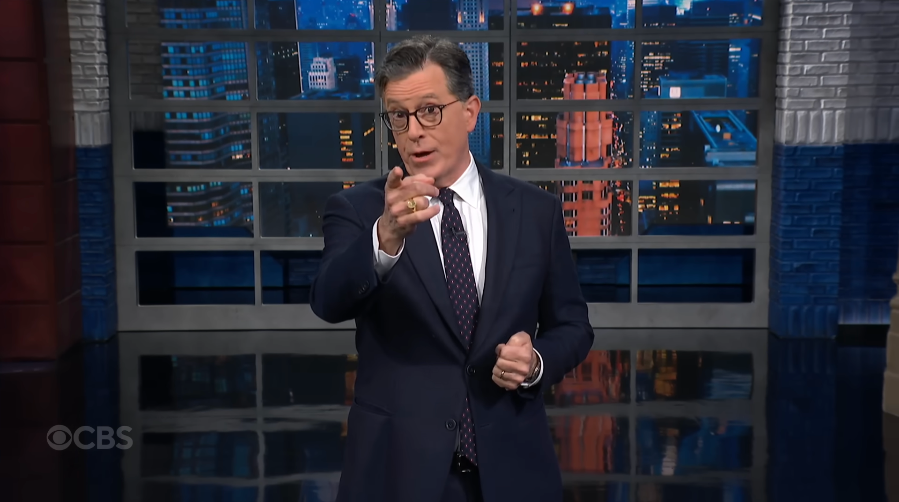 A person in a suit and tie stands on a set with cityscape backdrop, gesturing as if speaking to an audience
