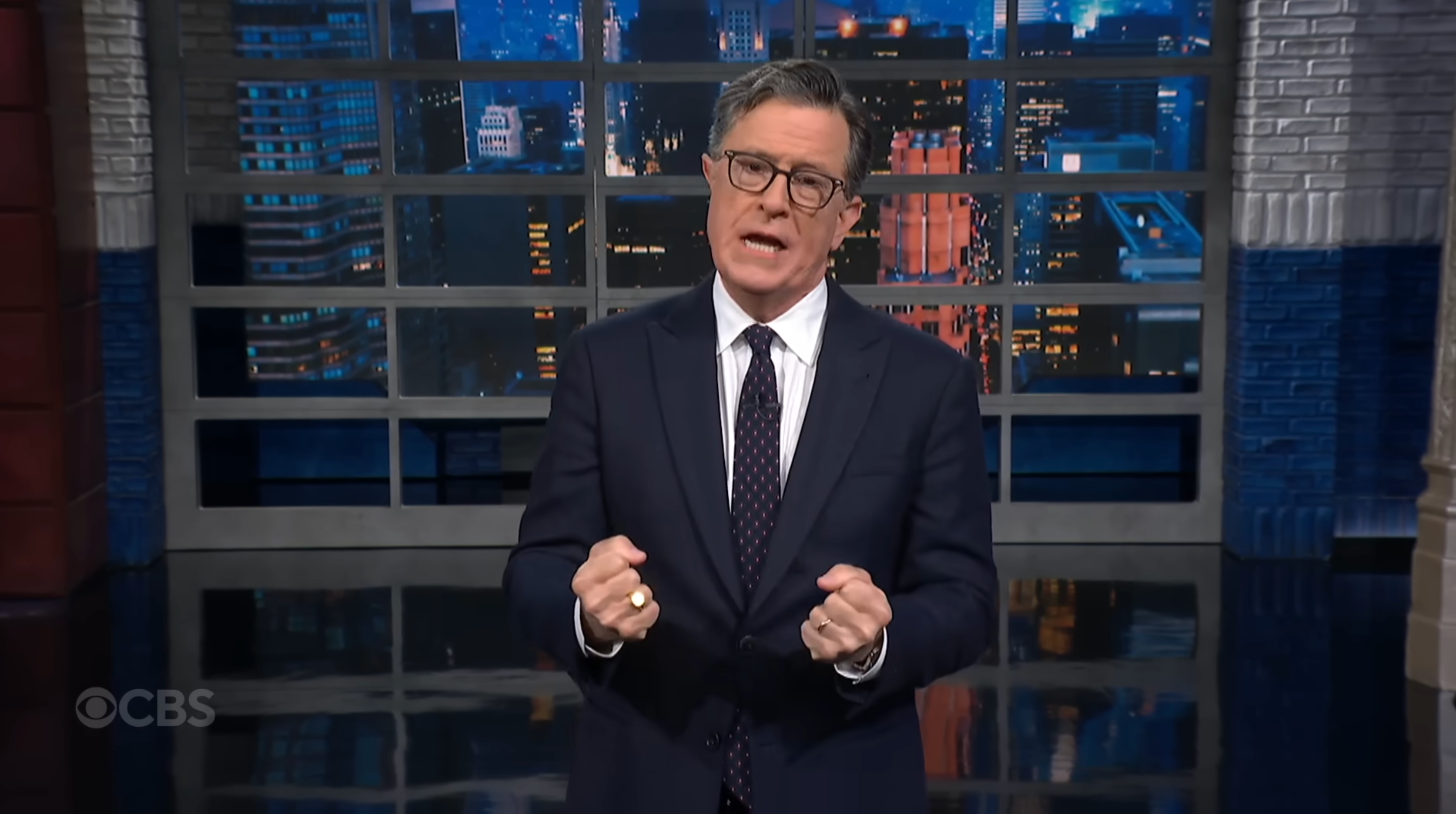 A person in a suit and glasses stands indoors, passionately speaking on a TV show set with a cityscape backdrop