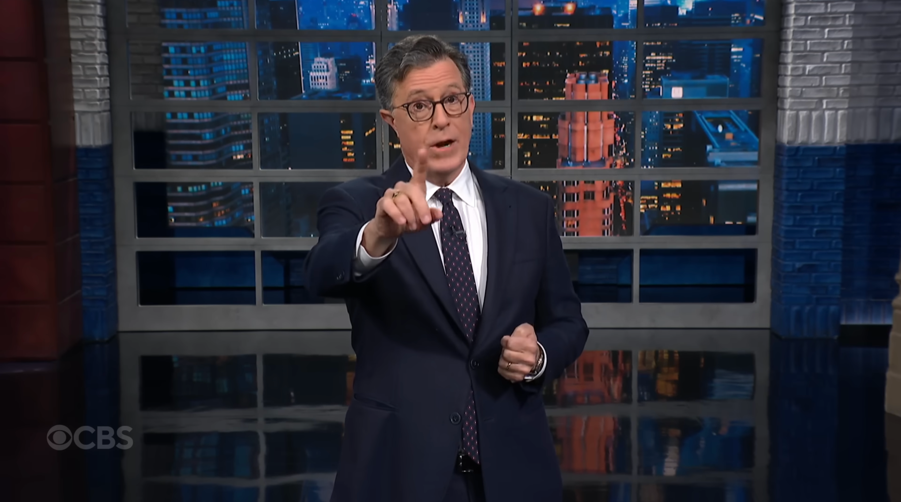 A person in a suit gestures forward on a television set with a cityscape backdrop