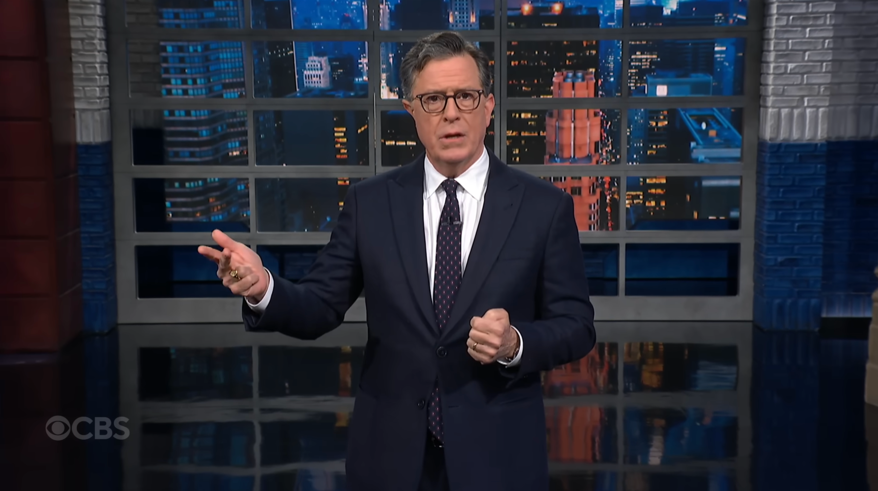A man in a suit and tie is hosting a television show, gesturing with one hand while delivering a speech in front of a cityscape backdrop