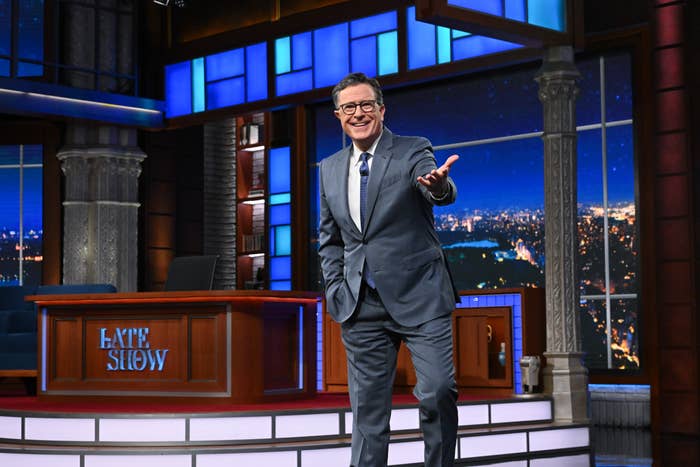Host on a late-night show set, wearing a suit, gestures warmly towards the audience