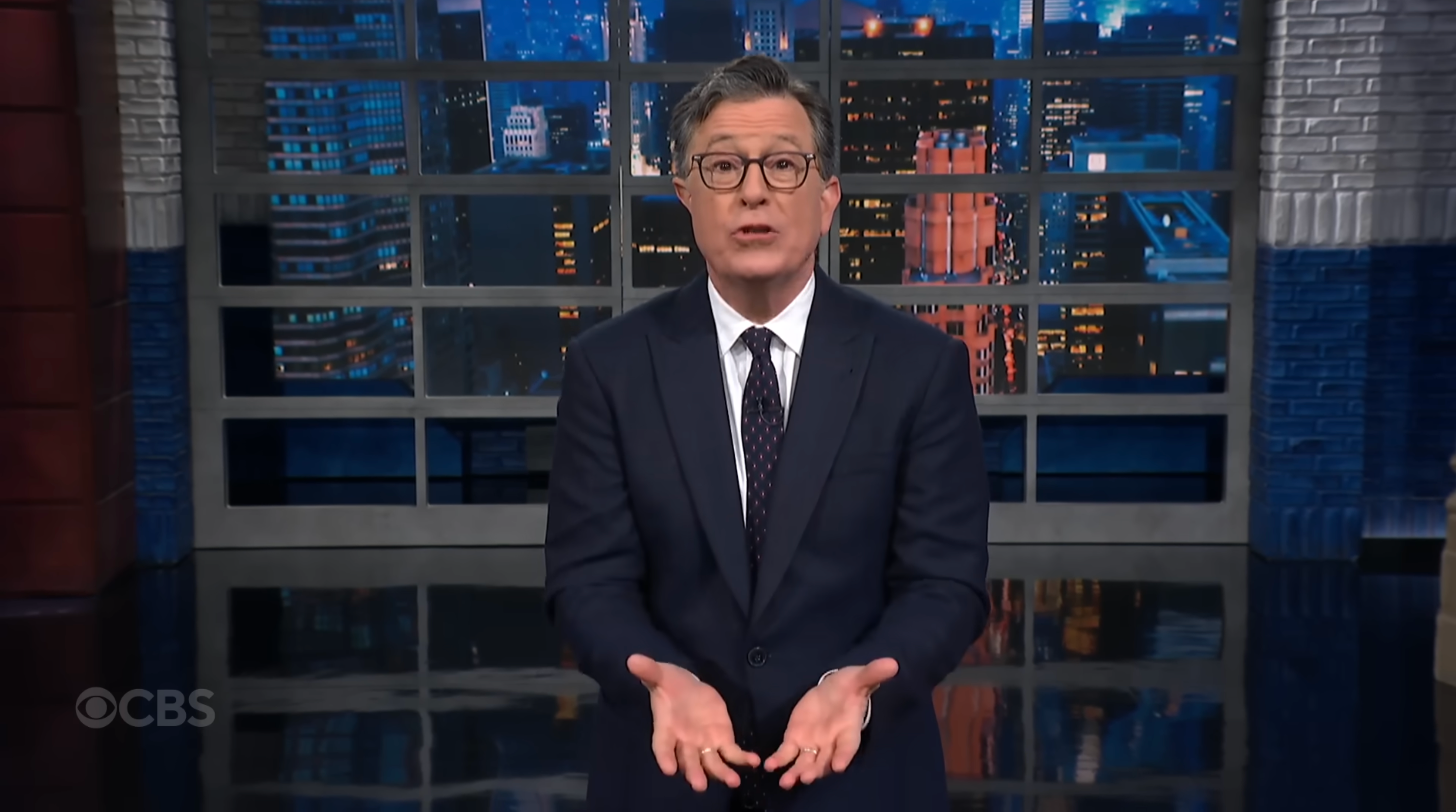 A person in a suit and glasses stands on a talk show set, gesturing with open hands during a monologue