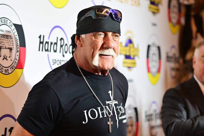 Person in a bandana and sunglasses at an event, wearing a T-shirt with a cross and text, standing on a logo-adorned backdrop