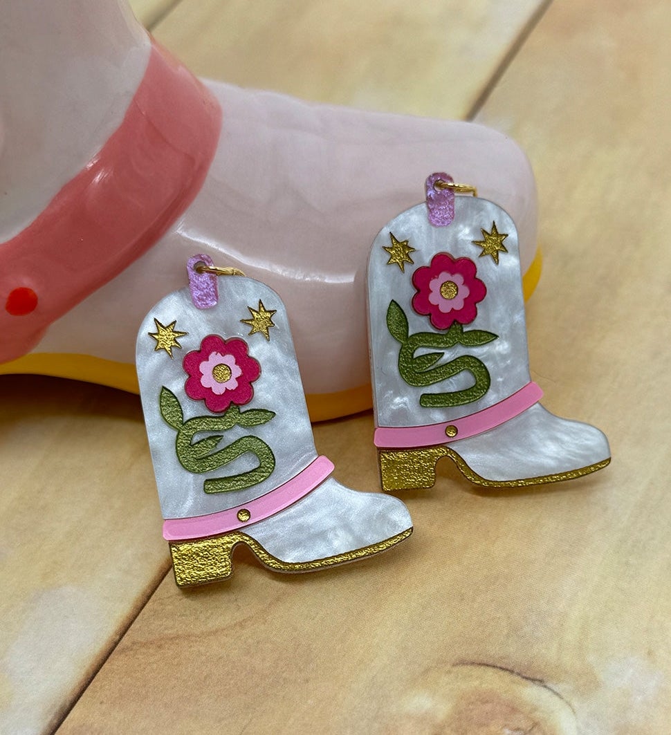 Cowboy boot-shaped earrings with a floral and snake design, resting on a wooden surface near a model foot