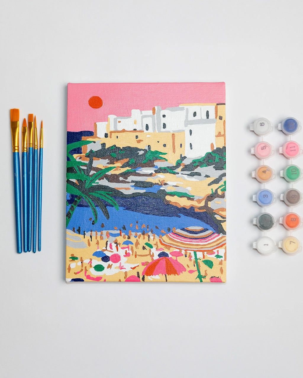 Paint-by-numbers canvas kit with brushes and paint pots, featuring an Italian coastal scene with umbrellas and buildings