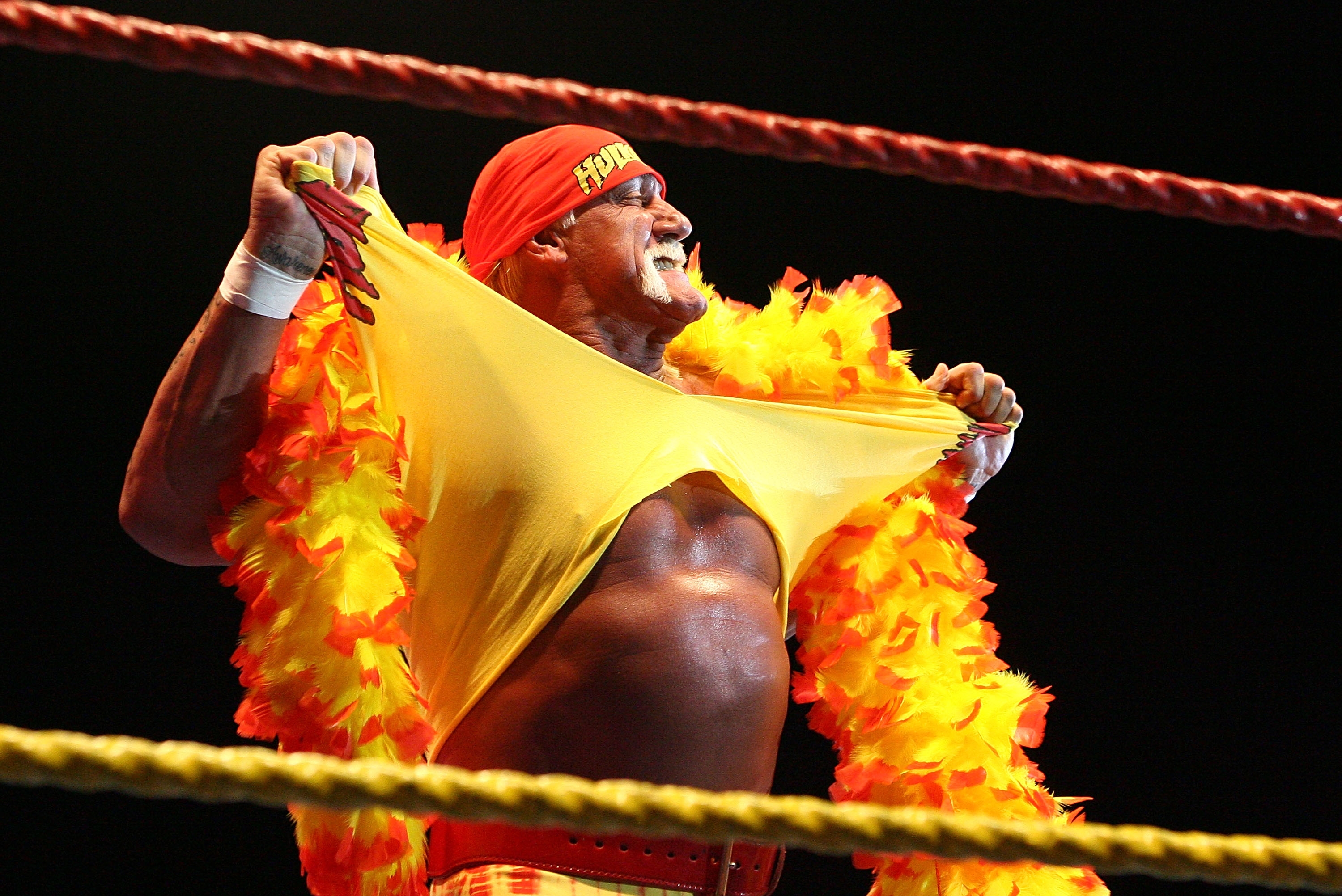 Wrestler in the ring tears a yellow shirt, wearing a red bandana and vibrant feather boa, exuding energy and charisma