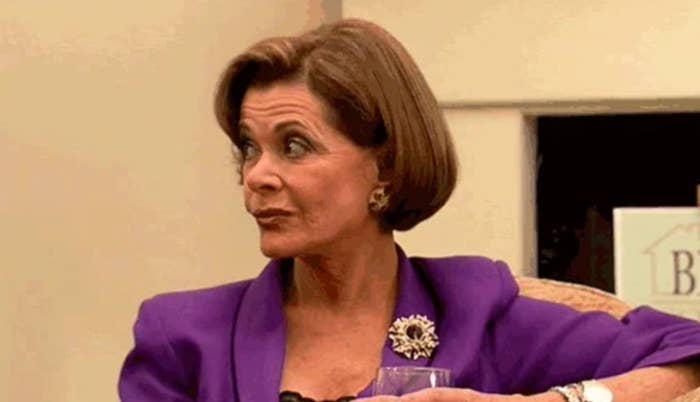 Woman with a bob haircut in a blazer, holding a drink and looking to the side with a raised eyebrow