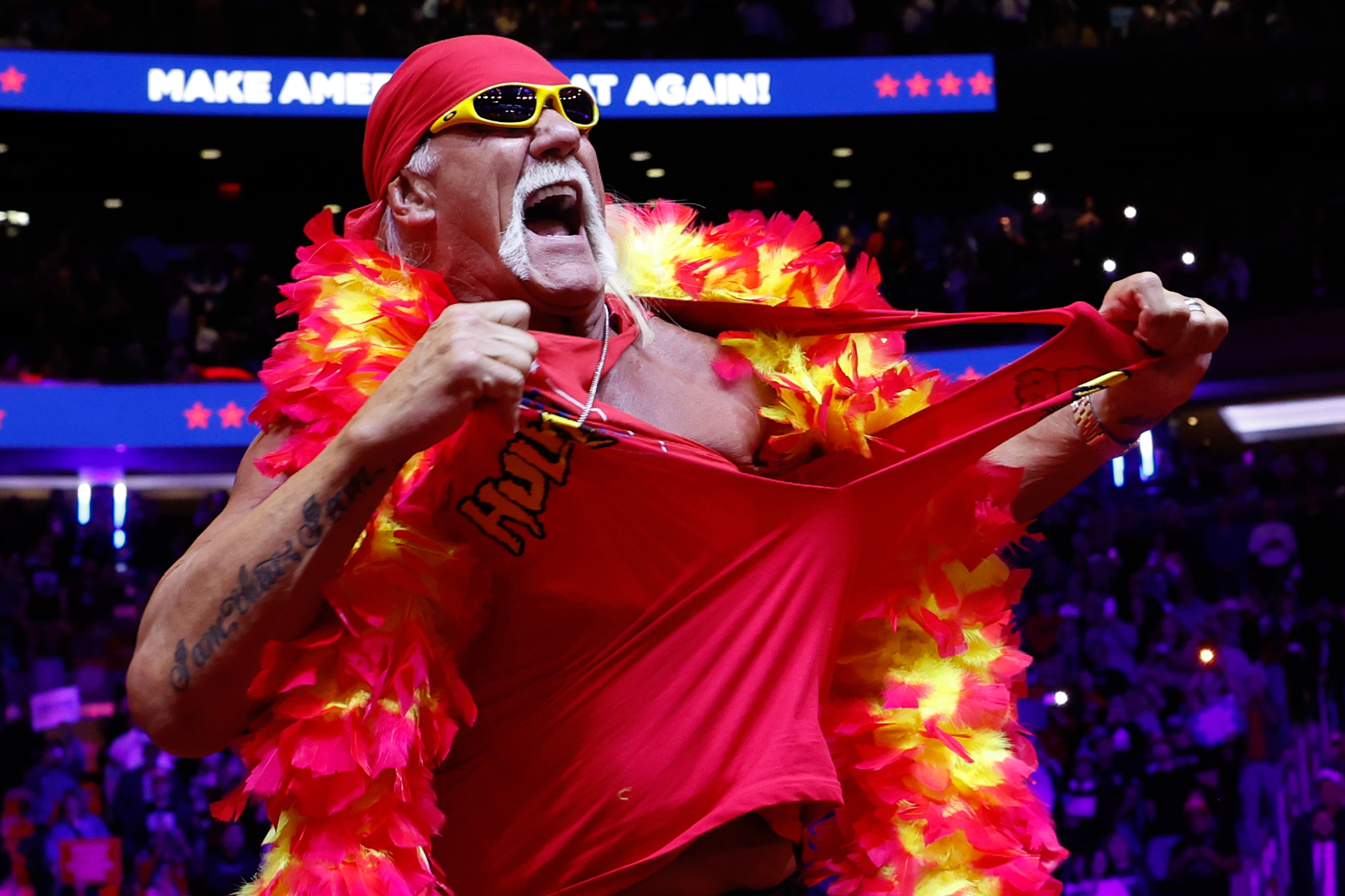 Hulk Hogan wearing feather boa, sunglasses, and bandana, tearing red shirt on stage with "Make America Great Again" sign in background at event